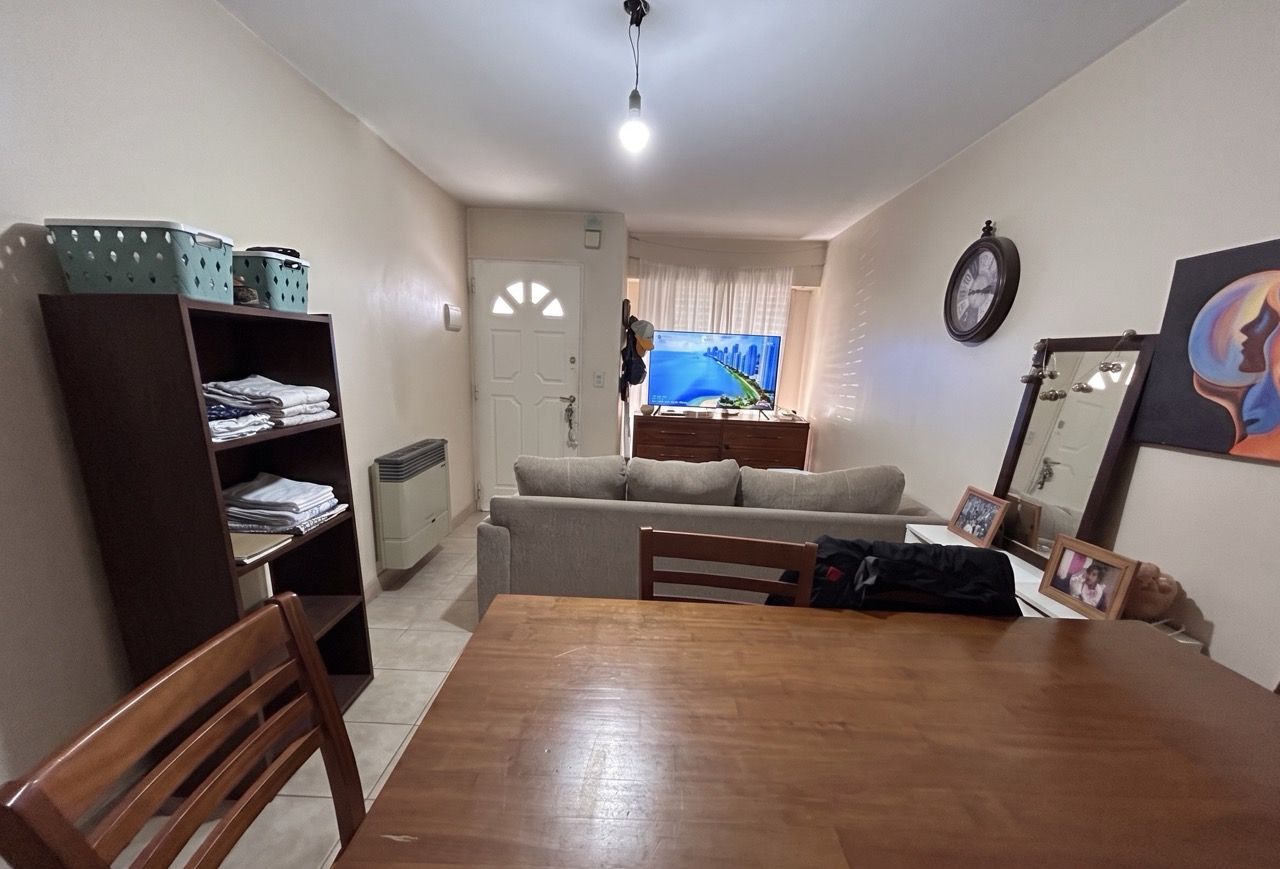 Living y comedor con sillón gris, mesa de madera y la puerta de entrada al fondo